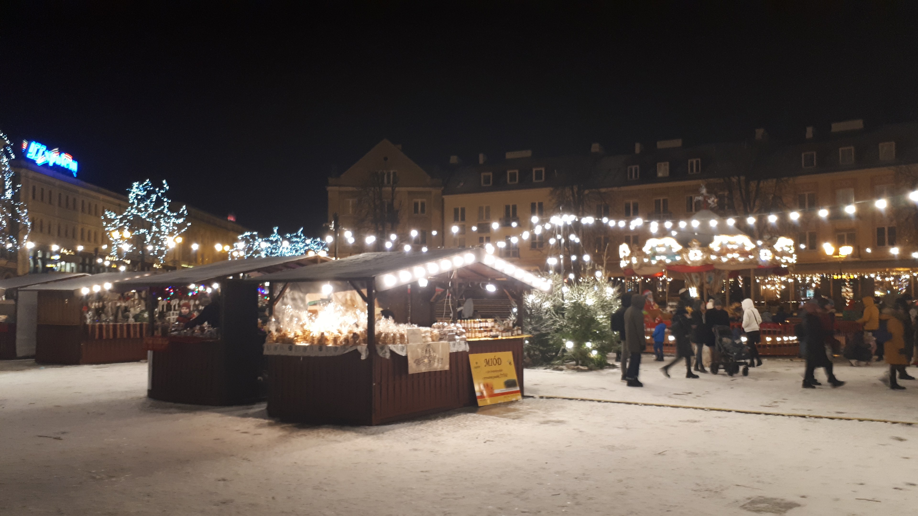 Ein geschäftiger Weihnachtsmarkt an einem verschneiten Abend mit Menschen, Buden, Pflanzen, Bäumen, Gebäuden und Schildern unter einem bewölkten Himmel.