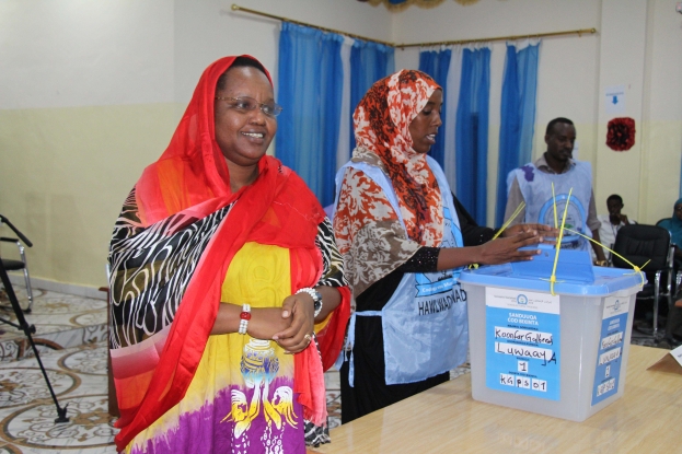 Zwei Frauen stehen nebeneinander an einem Abstimmungstisch mit Papieren und einer Box, während Menschen im Hintergrund unter einem Licht und einem Ventilator sitzen.