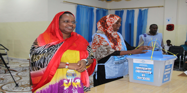Zwei Frauen stehen nebeneinander an einem Abstimmungstisch mit Papieren und einer Box, während Menschen im Hintergrund unter einem Licht und einem Ventilator sitzen.