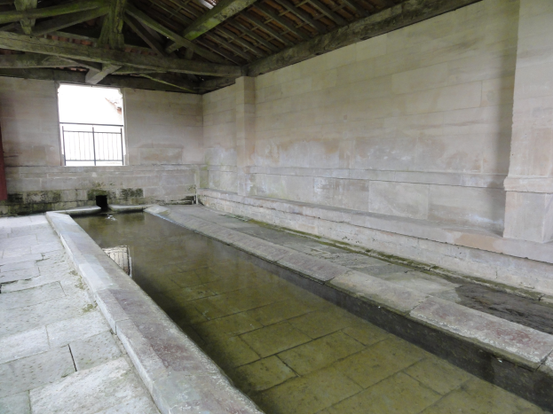Alter Steinbau mit einem zentralen Wasserbecken, einem Fenster im Hintergrund und hölzernen Balken oben, die an ein Bad erinnern.