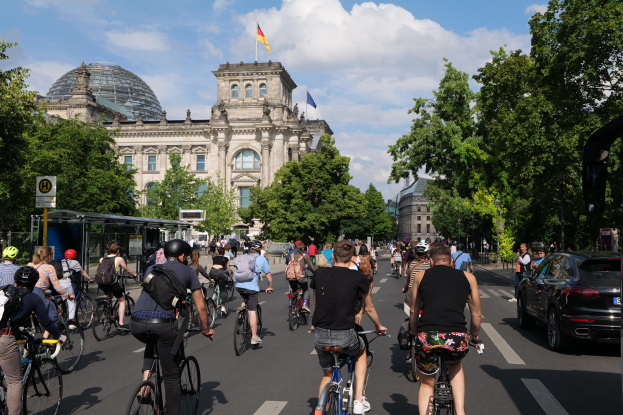 Eine Gruppe von Menschen, die auf Fahrrädern eine von Bäumen gesäumte Straße in Berlin, Deutschland, entlangfahren, mit Gebäuden und einem Bushaltestellen-Schild im Hintergrund, unter einem bewölkten Himmel und einer Flagge auf einem der Gebäude.