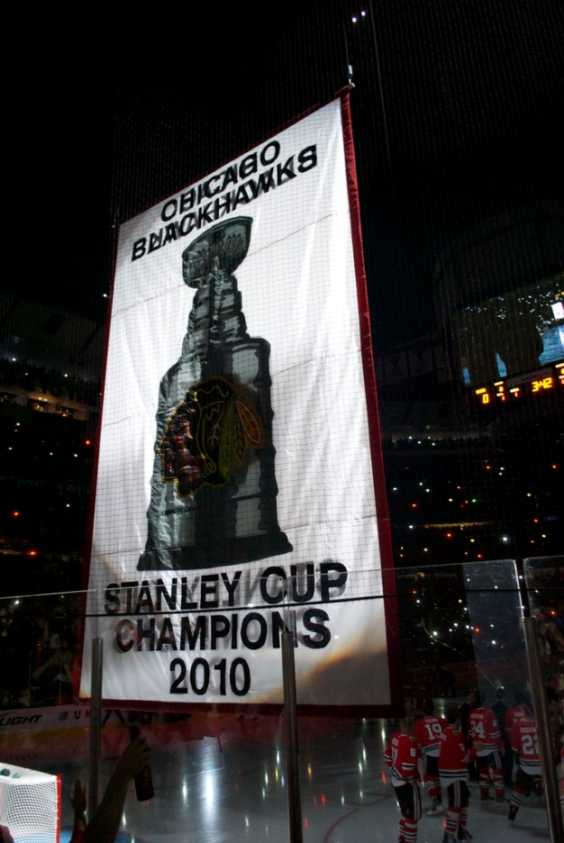 Eine nächtliche Stadionszene mit einem großen Banner in der Mitte, Zuschauern im Hintergrund, einem Glas auf dem Boden, einigen Spielern auf dem Feld und beleuchteten Rängen.
