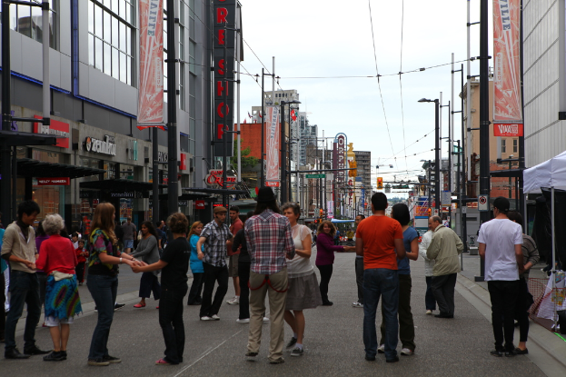Eine Gruppe von Menschen hält sich an den Händen auf einer Straße, die von Gebäuden mit Geschäften im Erdgeschoss, Straßenlaternen und Masten mit Drähten und Werbetafeln gesäumt ist; Zelte sind auf dem rechten Gehweg sichtbar.