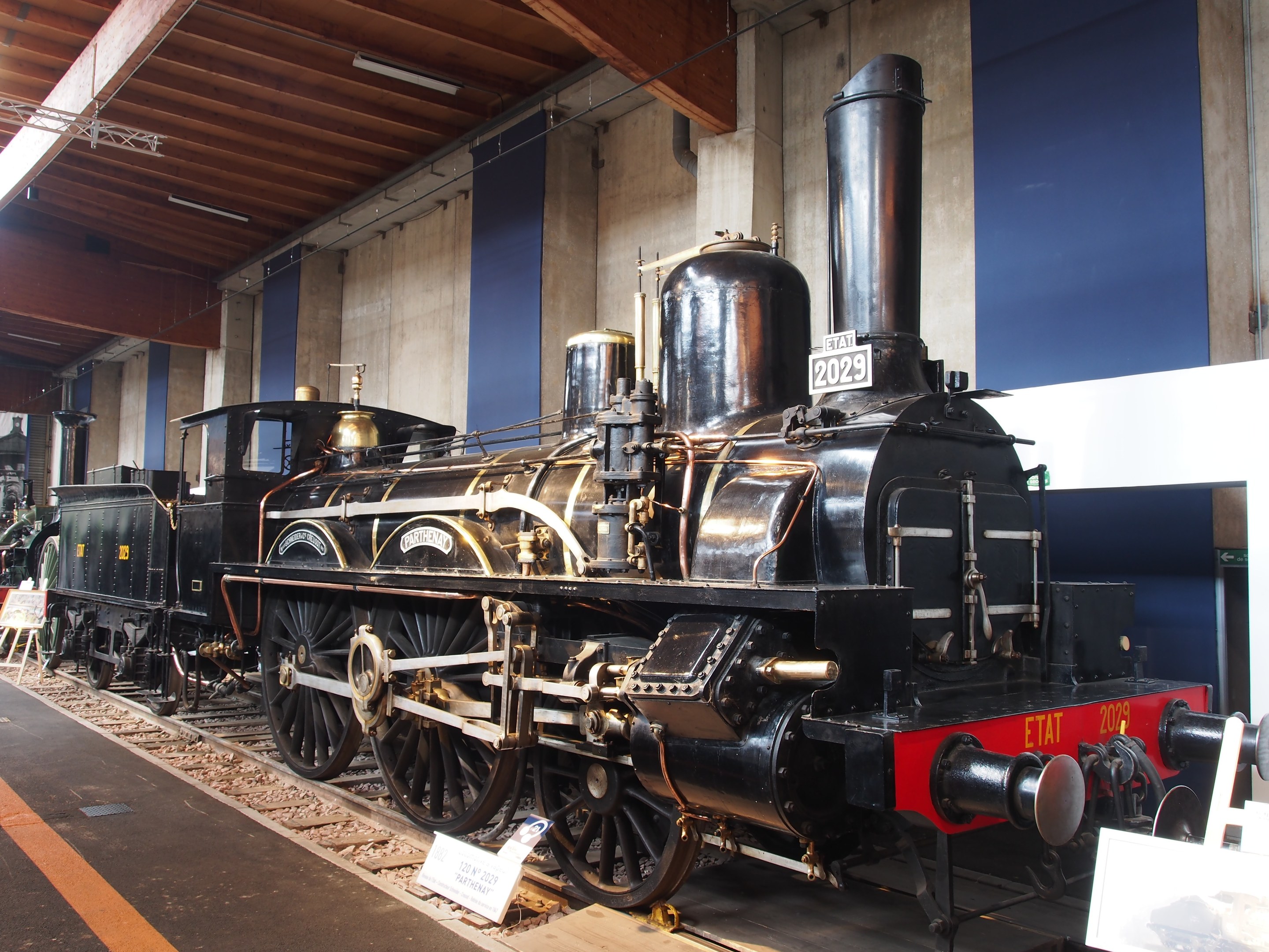 Eine große, alte Dampflokomotive in einem Museums-Setting umgeben von Eisenbahnschienen, Informations-Tafeln, Ständen und anderen Ausstellungsstücken, mit einer beleuchteten Decke und Wand im Hintergrund.