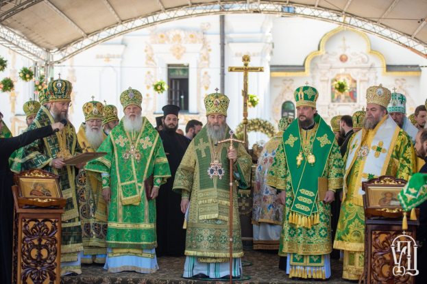 Eine Gruppe von Priestern vor einer Kirche während einer religiösen Zeremonie, wobei einer ein Buch und ein Mikrofon hält, und ein Kreuzsymbol, Blumen und ein Gebäude im Hintergrund.