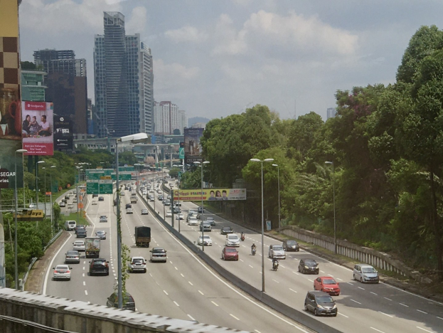 Eine vielbefahrene Autobahn mit hohen Gebäuden, Straßenlaternen, Schildern, Bäumen und einem bewölkten Himmel.