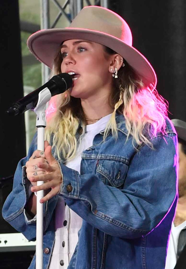 Miley Cyrus performs bei der CMA Music Festival in Nashville, Tennessee, mit Hut und Mikrofon auf einem Ständer, neben einer Person mit Mütze und verschiedenen Musikinstrumenten im Hintergrund.