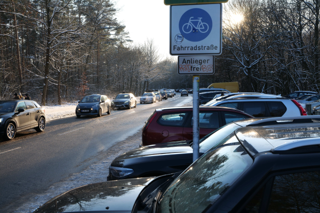 Ein Stau auf einer verschneiten Straße mit parkenden Autos, ein Mastschild im Vordergrund, Tänne und einen klaren Himmel im Hintergrund.