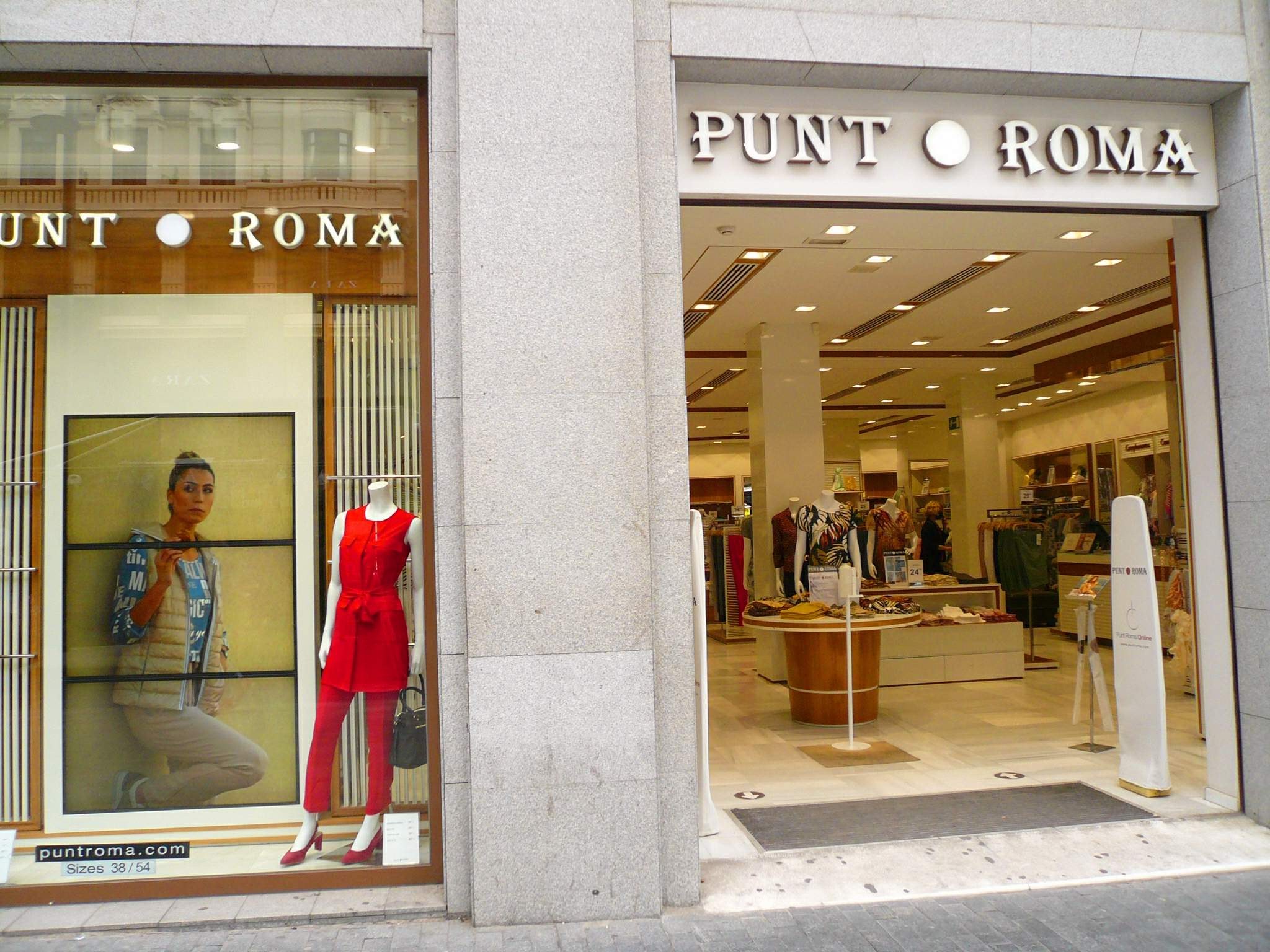 Vitrine des Punt Roma in Mailand, Italien, mit Schaufensterpuppen in Kleidern, beleuchtet von Deckenlampen, mit Text an der Wand und einem Türmatten auf dem Boden.