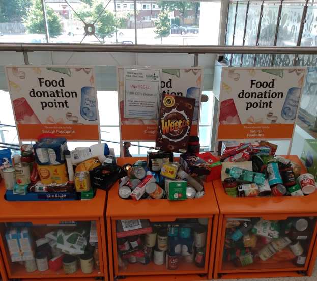 Orangene Schränke mit Lebensmittelspenden gefüllt, einschließlich Flaschen, Dosen und Kartons, mit Informationsschildern in der Nähe, durch Glasfenster betrachtet, die Außenfahrzeuge, Bäume, Gebäude und Himmel zeigen.