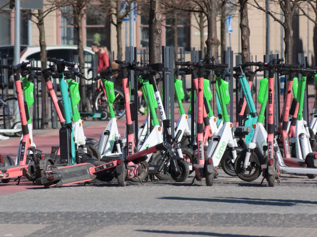 Eine Reihe von Elektro-Rollern, die an einer Straße geparkt sind, mit einer Person im Vordergrund und Baumwipfeln und Gebäuden im Hintergrund.