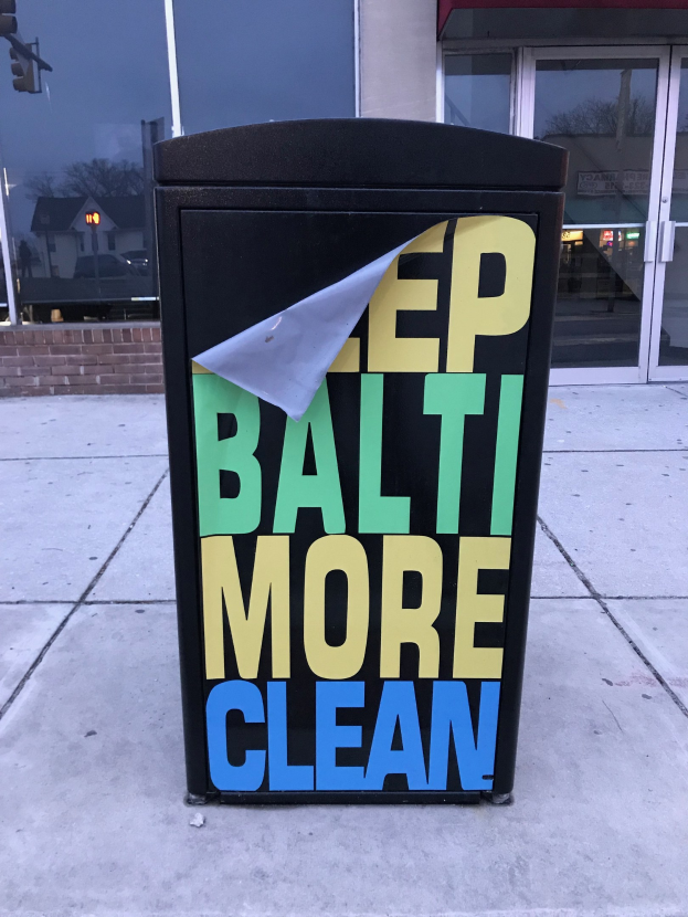 Schwarzer Müllcontainer mit "Keep Baltimore More Clean"-Text auf dem Gehweg vor einem Gebäude mit Glastüren.