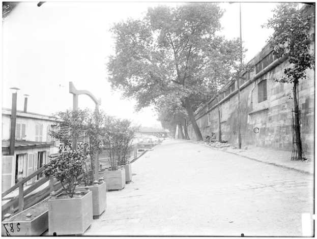 Ein Schwarz-Weiß-Foto einer Straße mit einer Wand auf der rechten Seite, Bäumen und Pflanzen auf der linken Seite und einer Brücke im Hintergrund, mit Text unten, der besagt: "New York City, New York, New Jersey, NY, USA, USA - Straßenansicht des New Yorker Staatsgefängnisses".