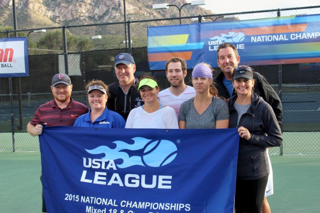 Gruppe von Menschen auf einem Tennisplatz mit einem "2015 USTA National Championships"-Banner, einer Umz├Ąunung, Bannern, Laternenmasten, B├Ąumen, Bergen und einem klaren blauen Himmel im Hintergrund.