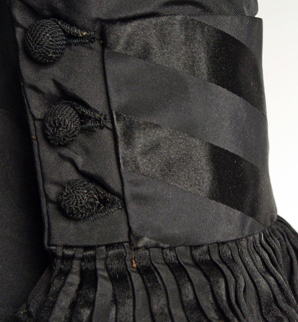 Nahaufnahme eines schwarzen, knielangen Kleides mit silbernen Knöpfen, die symmetrisch auf einem eng anliegenden Oberteil angeordnet sind, mit einem hohen Taillenbund, einem Rockschoß und leichtem Stoff vor einem weißen Hintergrund.