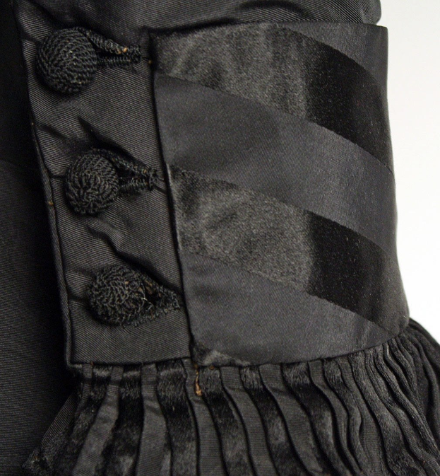 Nahaufnahme eines schwarzen, knielangen Kleides mit silbernen Knöpfen, die symmetrisch auf einem eng anliegenden Oberteil angeordnet sind, mit einem hohen Taillenbund, einem Rockschoß und leichtem Stoff vor einem weißen Hintergrund.