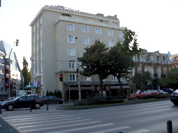 Eine befahrene Straße mit fahrenden Fahrzeugen, einer grünen Mittelleitplanke, Verkehrssignalmasten, Straßenlaternen, Bäumen, Gebäuden im Hintergrund und einem Himmel darüber.