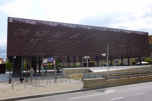 Großes Gebäude mit zahlreichen Fenstern, das den neuen Eingang zur Stadt bildet, umgeben von Straßeninfrastruktur, Fahrzeugen, Passanten, Bäumen und einem bewölkten Himmel.