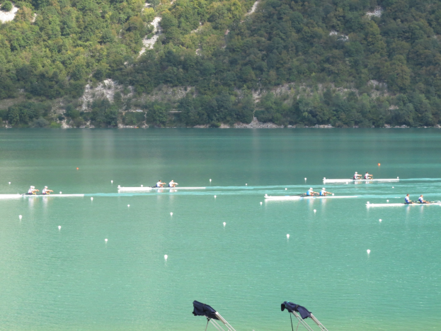 Gruppe von Menschen beim Ruderwettbewerb auf einem Gewässer mit Bäumen und einem Hügel im Hintergrund, Kleidung über Stühle am Boden drapiert.