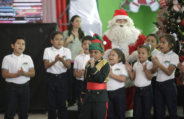 Kinder klatschen neben Santa Claus vor einem geschmückten Weihnachtsbaum, einer hält ein Mikrofon, mit einer Frau, Bildschirm, Vorhang und Wand im Hintergrund.