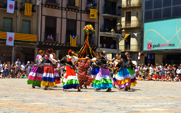 Eine Gruppe von Frauen in bunten Kleidern, die auf der Straße tanzen, umgeben von einer Menge Menschen, mit Gebäuden, Geländern, Balkonen, Fahnen, einem Laternenmast und einer Tafel mit Schrift im Hintergrund.