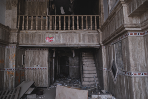 Ein altes Gebäudeinnere mit Schutt auf dem Boden, darunter hölzerne Gegenstände, Treppen, Geländer, Wände, Fenster und andere Gegenstände.