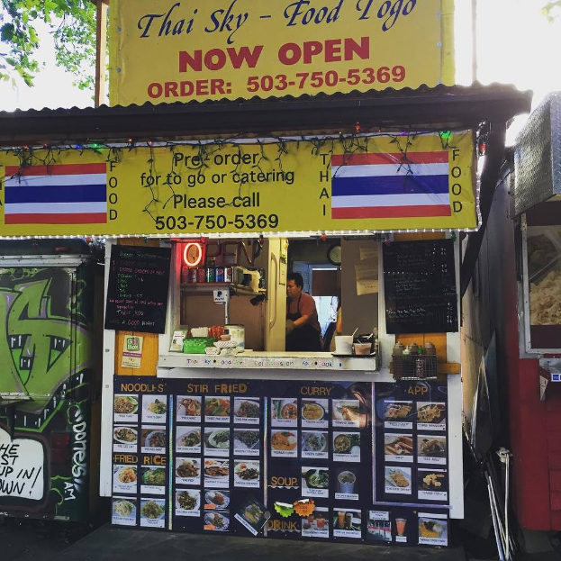 Ein Schaufenster mit verschiedenen Gegenständen wie Getränkedosen, Speisekarten, einer Uhr, Gewürzen in Plastikbehältern, Kondimenten, Werbetafeln und Namensschildern unter einem Himmel.