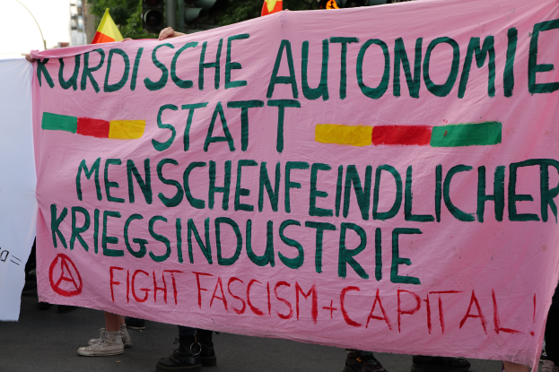 Eine Gruppe von Menschen steht auf einer Straße und hält ein pinkes Banner mit der Aufschrift "Kurdische Autonomie Statt Menschenfeindlicher Kriegsindustrie Fight Fascism Capital", mit Verkehrsampeln, Flaggen, Bäumen, Gebäuden und einem klaren blauen Himmel im Hintergrund.