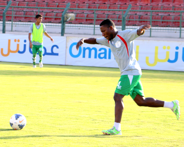 Ein Mann kickt einen Fußball auf einem Rasenfeld in einem Stadion mit zwei Personen in der Nähe und Bannern im Hintergrund.