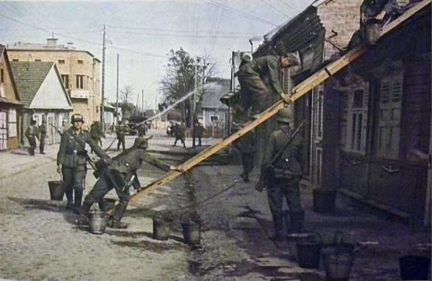 Eine Gruppe von Männern in deutschen Militäruniformen aus dem Zweiten Weltkrieg steht auf einer Straße neben einem Gebäude, trägt Helme und hält hölzerne Gegenstände, mit Eimern auf dem Boden, und Gebäuden, Bäumen, Polen, Drähten und einem klaren Himmel im Hintergrund.