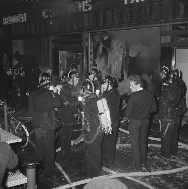 Schwarzes und weißes Bild von Feuerwehrleuten in Uniform mit Atmungszylindern, die neben einem Fahrrad und einem Tisch mit Gegenständen stehen.