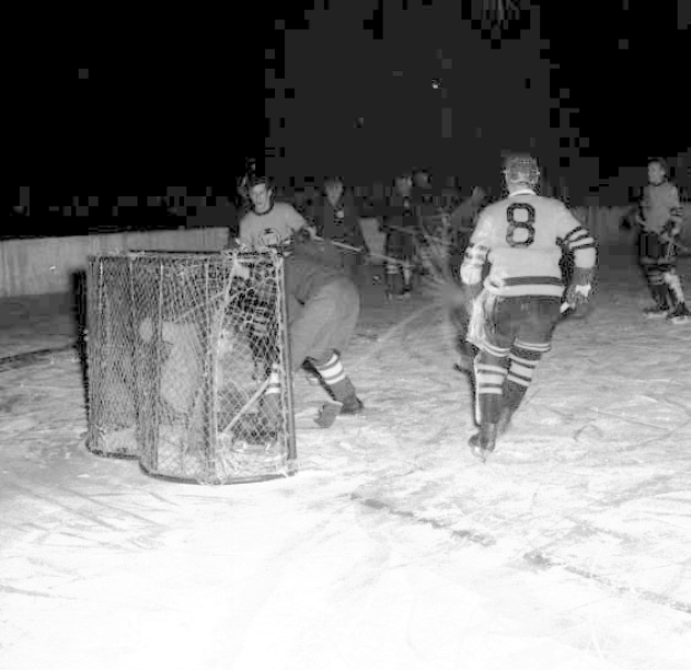 Schwarzes und weißes Foto von Männern, die Hockey auf einem Eisstadion mit einem Netz im Vordergrund und einer Wand im Hintergrund spielen.