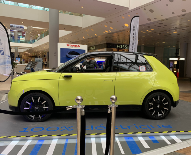 Ein gelbes wasserstoffbetriebenes Elektroauto vor einem Gebäude geparkt, umgeben von Absperrpollern mit Seilen, Bannern und Schildern, mit einer Person im Auto, einer Gruppe von Menschen in der Nähe, einer Säule, einem Topf mit einer Pflanze, Deckenleuchten und einer Decke mit Deckenventilatoren.