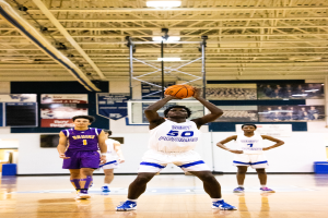 Ein Mann in einem Turnsaal, der einen Basketball schießt, mit Menschen im Hintergrund in Sportkleidung und Turnschuhen, unter einer Decke aus Stangen und Lampen.