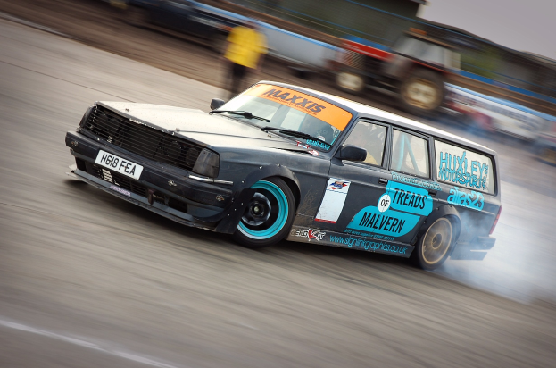 Ein Auto rast eine Rennstrecke entlang und hinterlässt eine Rauchspur, umgeben von anderen Fahrzeugen und Zuschauern.