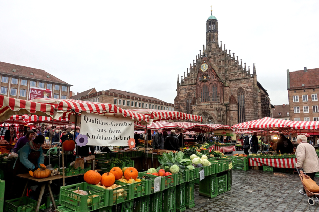 Ein belebter Outdoor-Markt in Nürnberg, Deutschland, der verschiedene Früchte und Gemüse anbietet, mit Menschen, die einkaufen und Zelte aufbauen, vor einem Hintergrund aus Gebäuden und einem Uhrenturm unter einem sichtbaren Himmel.