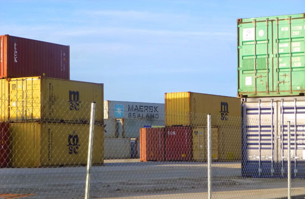 Ein hoher Stapel von Containern hinter Maschendraht mit einer Straße darunter und einem bewölkten Himmel im Hintergrund.