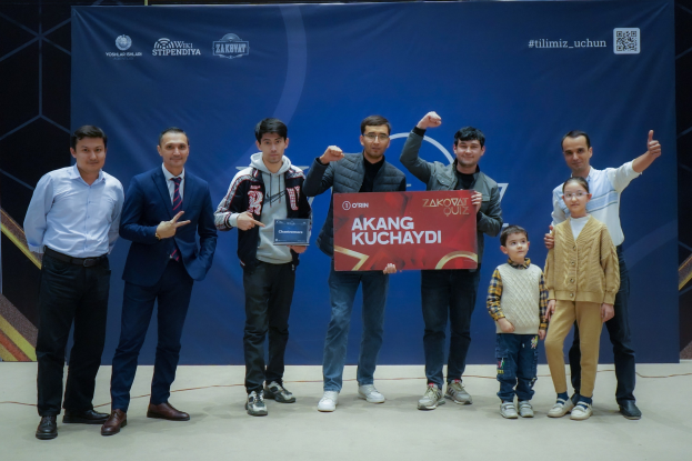 Eine Gruppe von Menschen auf einer Bühne, die eine Tafel mit dem Text "Akang Kuchaydi - Zakovat Quiz" halten, mit einem Banner, Scheinwerfern und anderen Gegenständen im Hintergrund.