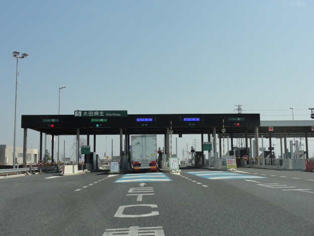 Eine Mautstation mit einem vor ihr geparkten Lastwagen, neben einer Straße mit weißen und blauen Linien, Laternenmasten, Schildern und anderen Gegenständen auf beiden Seiten. Im Hintergrund sind Gebäude, Bäume und ein klarer Himmel zu sehen.