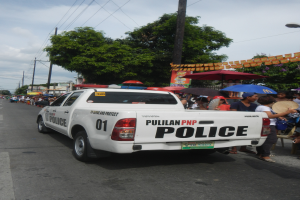 Ein Polizeiwagen fährt auf einer Straße mit einer Gruppe von Menschen auf der Seite, einige halten Schirme, mit Bäumen, Polen, Drähten, Gebäuden und einem klaren blauen Himmel im Hintergrund.