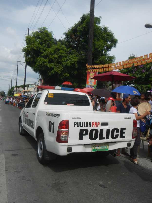 Ein Polizeiwagen fährt auf einer Straße mit einer Gruppe von Menschen auf der Seite, einige halten Schirme, mit Bäumen, Polen, Drähten, Gebäuden und einem klaren blauen Himmel im Hintergrund.