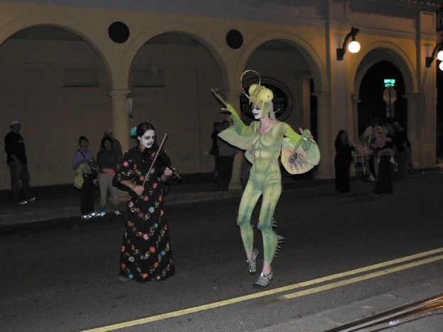 Eine Person in einem Kostüm steht neben einer Frau, die eine Geige spielt, mit Menschen und einem Gebäude im Hintergrund und Lichtern an der Wand, mit einer Straße unten.