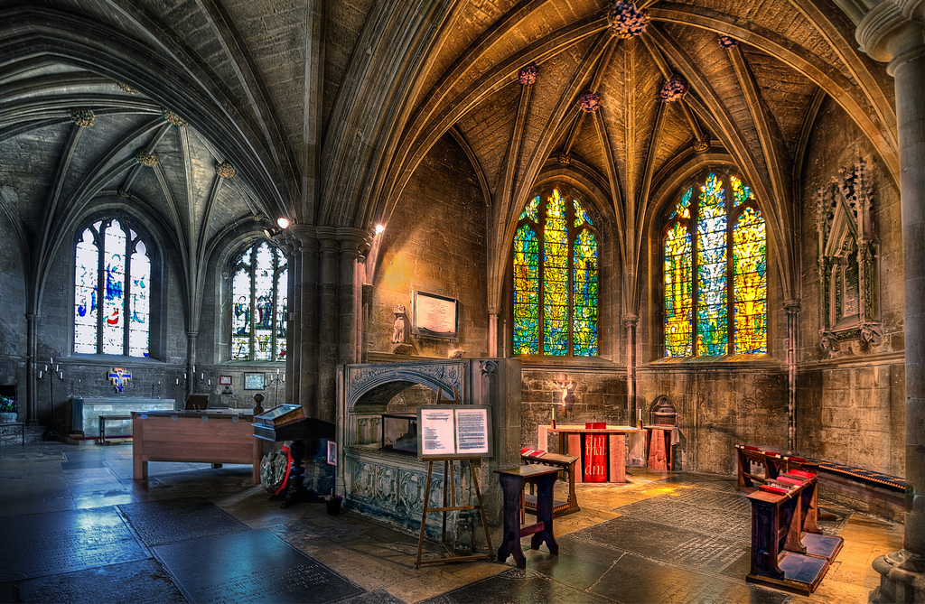 Ein Raum mit Bänken, Tischen, einer Tafel, Fenstern, einer Wand, Kerzen, einem Kamin und Lichtern in der Mitte, digital bearbeitet.