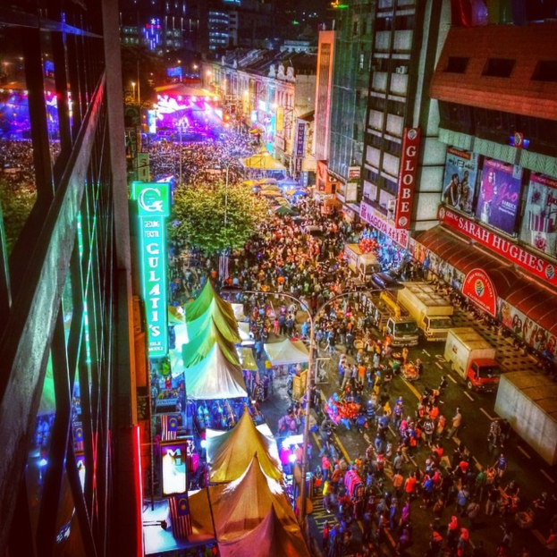 Ein buntes Karnevalszenario mit einer großen Menschenmenge auf der Straße, umgeben von Zelten, Geschäften, Gebäuden und Bannern, mit hell erleuchteten Bereichen im Hintergrund.