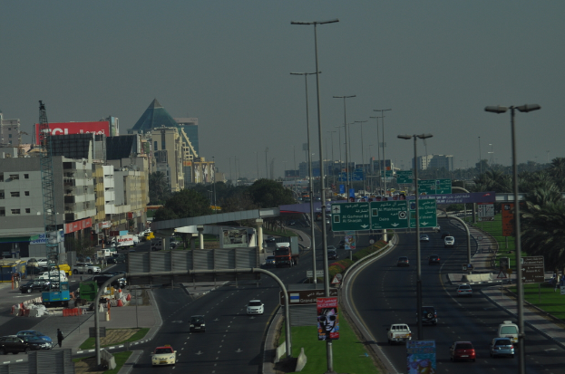 Eine Luftaufnahme einer Stadt mit Gebäuden, Türmen, Kraftfahrzeugen, Werbetafeln, Schildern, Straßen, Bäumen, Straßenlaternen, Himmel, Menschen, Absperrgittern und Informationsanzeigen.
