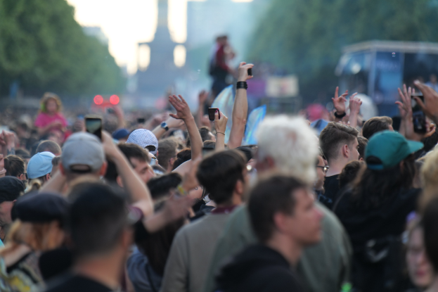 Eine Menge auf einem Musikfestival mit erhobenen Händen, einige tragen Mützen und halten Telefone, vor einem unscharfen Hintergrund aus Bäumen, Gebäuden, Lichtern und einem Fahrzeug.