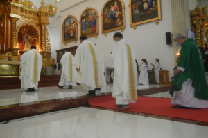Eine Gruppe von Priestern geht einen roten Teppich in einer Kirche entlang, während Menschen auf dem Boden vor und rechts von ihnen stehen und Fotoalben an der Hintergrundwand hängen.
