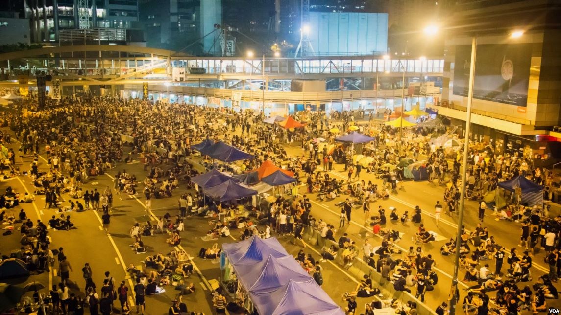 Eine große Menschenmenge steht auf einer nächtlichen Straßenprotest in Hong Kong, umgeben von Gebäuden mit Glaswänden, Zelten und einer Brücke im Hintergrund.