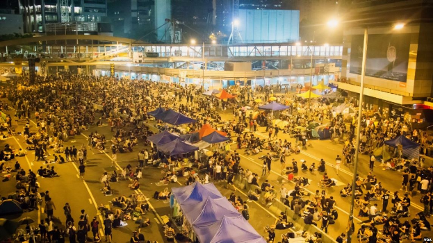 Eine große Menschenmenge steht auf einer nächtlichen Straßenprotest in Hong Kong, umgeben von Gebäuden mit Glaswänden, Zelten und einer Brücke im Hintergrund.