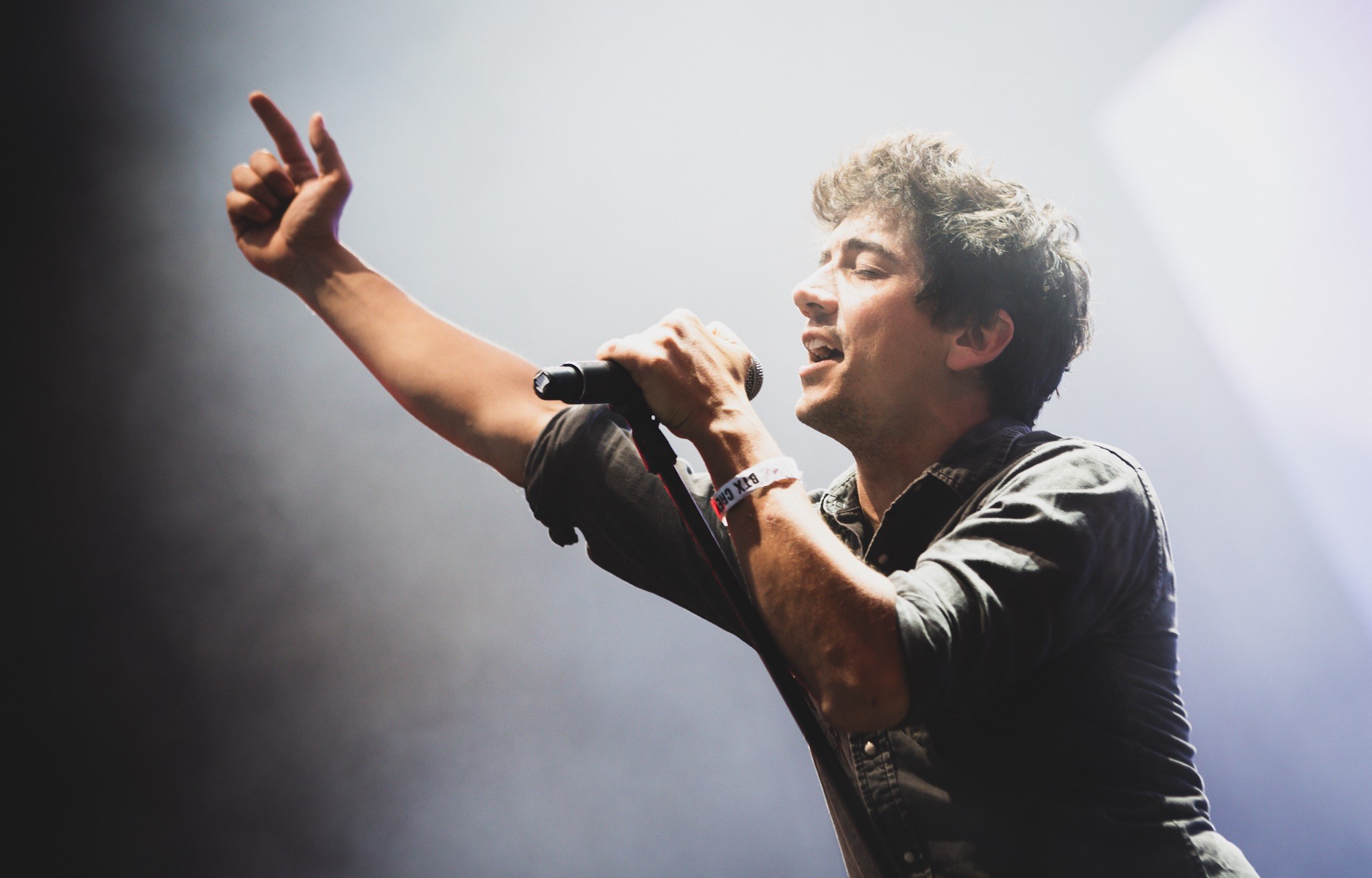 Ein Mann in schwarzem Hemd und Jeans singt leidenschaftlich in ein Mikrofon auf einer hell erleuchteten Bühne, mit einem engagierten Publikum, das im Hintergrund klatscht und jubelt.